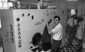 Tvättare, 28 maj 1966


En man klädd i vit skjorta och svarta byxor lyfter upp ett klädesplagg ur en låda. Han står invid en stor tvättmaskin. I bakgrunden står en kvinna klädd i vit arbetsrock intill en man även han klädd i vitt. Klädesplagg hänger på galgar i bakgrunden.
