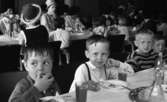 Inryckning, Gustavsson i Rynninge 24 maj 1966

Små barn sitter och äter vid dukade långbord i en matsal. I bakgrunden syns andra barn sitta vid andra långbord tillsammans med vuxna kvinnor klädda i klänningar. Damen som sitter närmast i bilden har en hatt på huvudet.