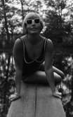 Baddräkter, Hus forts. 30 juni 1966

En fotomodell klädd i baddräkt och med solglasögon på sig poserar på en brygga vid ett vattendrag. Runt högra handleden har hon två armband.