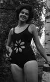 Baddräkter, Hus forts. 30 juni 1966

En fotomodell klädd i mörk baddräkt med en utskuren blomma i baddräktstyget på magen står och poserar invid ett träd. Hon håller ett par solglasögon i sin högra hand.