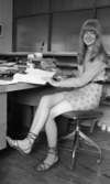 Kort- kort på jobb 2 augusti 1966

En ung kvinna med långt hår, långärmad tröja, kort kjol och med sandaler på fötterna sitter på en stol inne på ett kontor. Hon håller i en bok som ligger uppslagen framför henne på skrivbordet. En räknemaskin står också på skrivbordet.