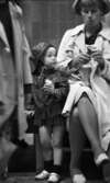 På stationen 1 16 augusti 1966

En liten flicka i kort kappa, kort klänning, strumpor, hatt på huvudet samt vita skor tittar intresserat på medan hennes mamma vecklar ut papperet på en chokladbit på Örebro järnvägsstation. Mamman är klädd i vit kappa, vit kort klänning, ljus hatt och vita skor.