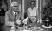 Missionärer i Kongo 29 augusti 1967

En äldre man klädd i ljus kostym, vit skjorta, mörk slips och som bär glasögon sitter vid ett bord och håller i en liten vit anteckningsbok med vänster hand. I sin högra hand håller han en penna. På bordet ligger en uppslagen tidning. Bredvid mannen sitter en äldre kvinna i en ärmlös, kort klänning på en stol.