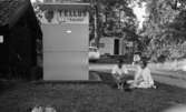 Missionsläger 10 augusti 1967

På ett missionsläger sitter en man och en äldre kvinna på en filt. Mannen är klädd i solglasögon, vit skjorta, ljusa byxor och randiga strumpor. Kvinnan är klädd i ljus, kort klänning, har en vit kofta över axlarna, vita sommarskor samt bär glasögon. På ett litet hus bredvid dem står en skylt där det står 