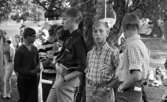 Missionsläger 10 augusti 1967

På ett missionsläger står tre unga pojkar vid ett träd. Pojken till vänster är klädd i en mörk skjorta med uppkavlade ärmar och jeans. Han håller en liten bok i sin högra hand. Pojken i mitten är klädd i rutig skjorta med uppkavlade ärmar och jeans och pojken till höger bär ock så rutig skjorta och jeans. Ytterligare ungdomar och tält syns i bakgrunden.