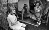 Missionsläger 10 augusti 1967

En vuxen kvinna sitter utanför ingången till ett tält med ett staket runt sammanfogat av pinnar tillsammans med fyra stycken ungdomar- tre flickor och en pojke. Hon har en afrikansk skulptur i sitt knä.