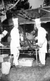 Natt 702, 12 juni 1967

Två män i kockhattar och arbetskläder håller griskött emellan sig som är uppspänt på en träbit. de håller på med att grilla kött. I bakgrunden syns en till kock som förbereder kött på en bänk. I bakgrunden står kvinnor och män och samtalar. 
Mannen till höger med kockmössa heter Evert Wing.