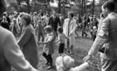 Midsommar, 26 juni 1967

Närbild på barn, kvinnor och män som dansar ringdans under midsommarfirande. Det är flera stycken ringar av dansande människor. En kvinna bär en vit kappa, flera män har kavajer och en man till höger har en kamera i en rem på axeln.