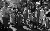 Midsommar, 26 juni 1967

Närbild på barn, kvinnor och män som dansar ringdans under midsommarfirande. Det är flera stycken ringar av dansande människor. En kvinna bär en vit kappa. En man till vänster håller i ett mikrofonstativ och har en cigarrett i munnen. En kvinna till höger är klädd i folkdräkt. Många män är klädda i kostymer.
