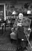 Kräftor- Karlslundsgård 5 aug. 1967

En äldre herre sitter i en fåtölj och håller i en käpp. Han är klädd i rutig skjorta, byxor, tofflor och rutig kavaj. Bakom honom syns bord, stolar, en soffa och en bonad på väggen.