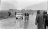 Orubricerad 18 maj 1967Under en cykeltävling kommer tre tävlingscyklister cyklande på en väg klädda i idrottskläder och med hjälmar på huvudet. En bil kör också på vägen med en skylt på sig där det står: 