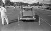 Orubricerad 21 augusti 1967

En man och en kvinna sitter i en Opel på en parkering. De är i färd med att köra iväg. En man i vit arbetsoverall står och pratar med en person till vänster. Ytterligare personer och bilar syns i bakgrunden. Även ett hus syns längre bort.