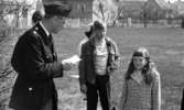 Oscaria test, Skolornas riksfinal, Smyckar holmens skola 12 maj 1967

Två pojkar i yngre tonåren, en tioårig flicka och en äldre dam står tillsammans med en polis på en gräsmatta. Den senare står i förgrunden och håller en papperlapp i sin högra hand. En av tonårspojkarna är klädd i jacka och byxor. Flickan har ljust, rutig kavaj och håret i pippilottor.