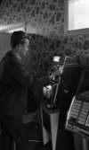 Knuttarna har bil 24 juli 1967

En tonårspojke drar i en spak tillhörande en spelmaskin inne i en fritidslokal. Han är klädd i mörk jacka, mörka byxor och ljus rutig skjorta. Det står flera flipperspel i lokalen.