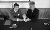 Parti ### (soc.), 18 augusti 1967
En man i rutig ljus kostym, Helge Hagberg, vit skjorta och mörk slips sitter vid ett bord och håller en ordförandeklubba i sin högra hand. Han har en ring på långfingret. Bredvid honom till höger sitter en yngre man i mörk kostym, ljus tröja och med ett armbandsur runt sin vänstra handled och en ring på vänster ringfinger. Framför dem ligger ett askfat med en tändsticka och en cigarett i. En tändsticksask och ett papper ligger invid fatet.