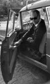 Konsum övar manöverkörning, 18 augusti 1967

En man sitter i framsätet på en bil klädd i mörk tröja, mörka byxor, vit skjorta och svarta solglasögon. Han befinner sig i centrala stan i Örebro.