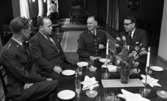 Kurator, Nye generalen 18 maj 1967

Inne i en elegant matsal sitter fyra herrar vid ett bord. Det är en ny general, en annan militär, landshövdingen Harald Aronson klädd i mörk kostym, vit skjorta och randig slips och en annan man klädd i mörk kostym, vit skjorta och svart slips. De sitter vid ett dukat bord. I bakgrunden står en kvinna i servitriskläder med ryggen mot kameran.