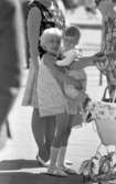 Kvar i stan 26 juli 1967

Närbild på ett syskonpar. En liten flicka i femårsåldern håller sin ettårsgamla bror i armarna. Hon är klädd i en kort, blommig ärmlös klänning, vita skor och vita strumpor. Brodern är klädd i långärmad vit tröja och rutig hängselbyxdress. Två vuxna kvinnor står nära barnen. En barnvagn står också bredvid barnen.