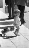 Kvar i stan 26 juli 1967

En liten flicka i tvåårsåldern drar på en rutig dockvagn där det sitter en docka. Hon är klädd i en långärmad, ljus blus, ljusa underbyxor, vita strumpor och vita skor. Hon går omkring i centrala stan i Örebro.