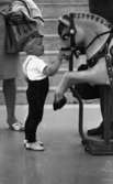 Kvar i stan 26 juli 1967

En liten pojke i tvåårsåldern står invid en stor leksakshäst och klappar den. Han är klädd i mörka hängselbyxor, vit kortärmad tröja, vita strumpor och vita skor. Han bär en mörk keps på huvudet. Bredvid honom står en kvinna i kort klänning, vita lågklackade skor och med en vit kofta över vänster arm. Hon har en stor handväska som också hänger över vänster arm.