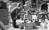 Kvar i stan 26 juli 1967

En kvinna i en kort ärmlös klänning sitter ned nära några lådor på ett torg i centrala Örebro. En kaffetermos står framför henne. En man står invid henne och böjer sig fram och talar med henne. Han är klädd i långärmad mörk tröja, mörka byxor, mörka strumpor och svarta skor. I bakgrunden står en bilar parkerade.