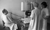 Lasarettet 14 juni 1967

En patient sitter på en stol med en klämma på näsan klädd i vit undersökningsrock. Han har ett rör i munnen med en slang som mynnar ut i fler slangar som är kopplade till något som ser ut som en syrgastub med en större mätapparat i anslutning till denna med stora pappersrullar på. Två sjuksköterskor klädda i vita rockar står med ryggarna mot kameran och betraktar resultatet på mätningsutrustningen.