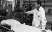 Lasarettet 14 juni 1967

En läkare klädd i vit läkarrock, vit skjorta och randig fluga står invid en operationssäng och håller sin vänstra hand på sängen och sin högra hand på undersökningsutrustning som står i bakgrunden.