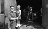 Hovslagaren tippar 26 augusti 1967

En far håller sin son i famnen. Fadern är klädd i rutig skjorta och mörka byxor med livrem. Sonen är klädd i vit T-tröja, mörka hängelbyxor, vita strumpor och vita träskor. De står framför en dörröppning vid en byggnad. Inne i byggnaden bakom dem står en bunden häst som lyfter högra hoven upp i luften.