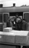 Frövi 2 juni 1967

Frövi järnvägsstation. Postleverans med tåg. En järnvägstjänsteman står i ett tåg med öppen dörr och håller två paket i famnen. Nedanför står en annan järnvägstjänsteman samt ytterligare två män klädda i rockar. Framför dem står en vagn med paket.