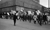Första maj demonstration 2 maj 1967

Ett första-majtåg går förbi varuhuset Krämaren i centrala Örebro. Många av deltagarna bär på skyltar med texter på. Banderoller förekommer också. Många män bär kavajer och byxor. Vissa är klädda i rockar. Kvinnorna är klädda i kappor.