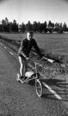 Glass, Nygren i Sten..., Gustavsvik 13 juni 1967

En man åker på en speciell sparkcykel på en landsväg. Han är klädd i mörk träningsjacka, mörka shorts, mörka strumpor och vita gymnastikskor. Sparkcykeln är utrustad med ett stort hjul framtill och två hjul baktill. Den har en låda i mitten avsedd för bagage. Landskapet syns i bakgrunden.