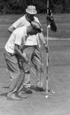 Grybe golf, Gustavsvik 17 juni 1967Två män klädda i vita skjortor och ljusa byxor spelar golf på en golfbana. Mannen i förgrunden bär en vit- och mörkrandig keps på huvudet och håller på och puttar ner den vita golfbollen med en klubba i ett intilliggande hål. Den andre mannen bär en vit- och mörkrutig hatt och står bredvid stången som en vimpel är fäst på och som står intill hålet. Han håller sin golfklubba i vänsterhanden.