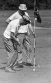 Grybe golf, Gustavsvik 17 juni 1967

Två män klädda i vita skjortor och ljusa byxor spelar golf på en golfbana. Mannen i förgrunden bär en vit- och mörkrandig keps på huvudet och håller på och puttar ner den vita golfbollen med en klubba i ett intilliggande hål. Den andre mannen bär en vit- och mörkrutig hatt och står bredvid stången som en vimpel är fäst på och som står intill hålet. Han håller sin golfklubba i vänsterhanden.