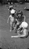 Grybe golf, Gustavsvik 17 juni 1967

I förgrunden syns två små pojkar i ettårsåldern som befinner sig i närheten av vattnet på utomhusbadet i Gustavsvik. Båda två har vita små mössor på sina huvuden och är i övrigt nakna. Pojken till vänster står upp i det grunda vattnet och håller sin vänstra hand på en tonårsflickas högra ben. Hon är klädd i en vit bikini och sitter på huk med en spade i sin högra hand. Hon skyfflar i sand i en liten leksakshink. Bredvid henne på andra sidan sitter den andre lille pojken i sanden och håller en leksaksspade i sin högra hand. Framför honom står en leksakshink. I bakgrunden syns bl.a. en kvinna i mörk baddräkt och en liten pojke i ljusa badbyxor.