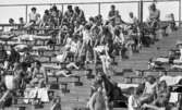 Gustavsvik 16 maj 1967

Personer i alla åldrar sitter på utomhusläktaren på Gustavsviks badhus. Många kvinnor är klädda i baddräkter och bikinis. Många män har badbyxor på sig. Flera personer sitter på handdukar.