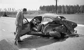 Götalundaolycka 29 maj 1967

I förgrunden syns en bil som varit med om en svår krock. Den är helt hoptryckt och står på vägen. En man klädd i grå kostym, vit skjorta och svart slips står vid sidan av bilen och blickar in i förarsätet. Framdörren på bilen står vidöppen. På andra sidan vägen i bakgrunden står en annan bil i diket och en person syns stå i närheten av denna.