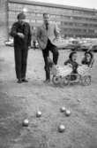 Med pappa på turné 14 april 1967

Den kände skådespelaren Helge Skoog spelar boule tillsammans med en annan man. Helges ettårsgamla barn sitter i en vit barnvagn med dubbelsits i närheten. Det ena barnet gråter. De befinner sig på parkeringen bakom Norlings bryggeri. Parkerade bilar syns även på bilden. Helge är klädd i vit kavaj, beige skjorta, svart slips, svarta byxor och svarta skor. Barnen är klädda i svarta dressar med svarta hattar på huvudena. Den andre mannen är klädd i svart jacka, randig skjorta, svarta byxor och svarta skor.
