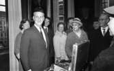 Mingporslin, Maranata, Strejken 25 oktober 1966

En äldre man står i förgrunden i ett rum med en lustig rund mössa på huvudet. Han är klädd i ljus kostym, vit skjorta och ljusrandig slips. Nedanför honom på ett bord står en stor resväska i silverskimrande metall fylld med mer mössor. Fler äldre herrar och damer syns i bakgrunden. En stor tavla föreställande ett porträtt av en man syns i bakgrunden.