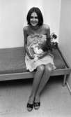 Miss annorlunda 10 oktober 1966

En fotomodell sitter på en säng och håller i en blombukett samt en skylt. Hon är klädd i en lårkort, vit klänning med en silverrosett framtill och ett snöre som löper från denna runt hennes hals. På fötterna har hon svarta lågklackade skor.