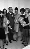 Miss annorlunda 10 oktober 1966

Fem fotomodeller står samlade tillsammans med en man som står i mitten klädd i ljusrandig kostym, vit skjorta och svart slips. Han håller en blombukett i sin högra hand. Modellerna är klädda i olika lårkorta klänningar och bär skyltar i sina händer. Varje fotomodell bär en skylt på med en siffra. Siffrorna på skyltarna är tal mellan 1-5.