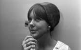 Modenummer 21 oktober 1966

Närbild på en ung kvinna som är modell. Hon har en hatt med hakrem på huvudet och bär ett ljust, mönstrat och ärmlöst plagg på överkroppen. Hon bär örhängen i form av stora vita pärlor i öronen och fyra ringar på höger ringfinger.