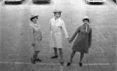Modenummer 18 april 1967

Tre fotomodeller står tillsammans på en bilparkering. Alla tre bär ljusa hattar med hakband runt hakan. Kvinnan till vänster har en ljus dräkt på sig och bruna knytskor. Kvinnan i mitten bär en vit kappa och bruna lågklackade pumps. Hon håller kvinnan till höger i handen. Denna bär en brun kappa och svarta, lågklackade skor. Bilar står parkerade i bakgrunden.
