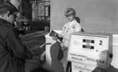 Bensinautomat 29 oktober 1966

en kvinna klädd i arbetskläder och med en tidning i handen står på en bensinstation. En man lyfter bensinslangen. Han är klädd i mörk rock och glasögon.