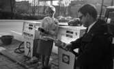 Bensinautomat 29 oktober 1966

På en Shellmack står en kvinna ur personalen klädd i arbetskläder och bläddrar i en tidning. En man står i förgrunden med en bensinpumpsslang i handen. Han är klädd i en svart rock, vitrutig skjorta, svart slips och bär glasögon. Bilar syns köra omkring på gatan i bakgrunden.