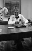 Lungkliniken, Nappivalen, Grekiskt handarbete 11 december 1967

En läkare på lungkliniken sitter vid bordet inne på sitt kontor och håller sina glasögon i händerna. Han är klädd i en vit läkarrock, vit skjorta och svart fluga. En blomvas står med en blomma till vänster i på en liten duk på bordet. Bakom doktorn hänger en tavla på väggen.