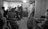Lungkliniken, Nappivalen, Grekiskt handarbete 11 december 1967

En modevisning inne i ett rum i ett hus. En fotomodell klädd i en ljus blus med trekvartslång ärm, en mönstrad kjol av grekiskt handarbete och svarta, lågklackade pumps står mitt på golvet. Publik sitter på stolar vid bord och betraktar henne.
