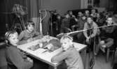 Lärare lärde film, Hantverksbutik, Hans Alsér, Ingenjörer på kurs 18 november 1967

Tre pojkar i tioårsåldern sitter vid ett vitt bord med hörlurar på sig samt under en mikrofon som hänger över deras huvuden. Det finns en till mikrofon invid pojken som sitter längst till höger. De har varsin läskedryck samt dricksglas framför sig på bordet. Deras klasskamrater sitter som publik på stolar i bakgrunden.