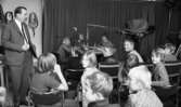 Lärare lärde film, Hantverksbutik, Hans Alsér, Ingenjörer på kurs 18 november 1967

Tre pojkar i tioårsåldern sitter vid ett vitt bord med hörlurar på sig samt under en mikrofon som hänger över deras huvuden. Det finns en till mikrofon invid pojken som sitter längst till vänster. De har varsin läskedryck samt dricksglas framför sig på bordet. Deras klasskamrater sitter som publik på stolar i närheten av dem. Till vänster står en medelålders herre som troligen är deras lärare.  Han är klädd i grå kostym, vit skjorta, svart slips och svarta skor.