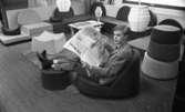Miss Backfisch Kandiden, Travtipset, Möbler på golvet 7 oktober 1967

En ung man klädd i ljusgrå kostym, vit skjorta och mörkrandig slips sitter i en mjuk möbel som är placerad på golvet och läser en tidning i ett rum.