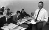 Mekanförbundet, 31 maj 1968

En grupp män sitter vid bord och skriver på Mekanförbundet. De sitter med ritningar och skriver på papper och gör olika matematiska beräkningar. De går en mekanikerutbildning. Tre män som sitter längst fram på bilden är klädda i kostymer, skjortor och slipsar. Framför dem står en lärare i mekanik och ler in i kameran. Han är klädd i vit skjorta, ljus slips samt ljusa byxor och håller ett papper i händerna.